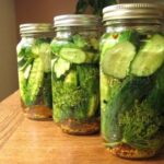 Three glass jars of cucumbers, dill, and spices on a wooden table